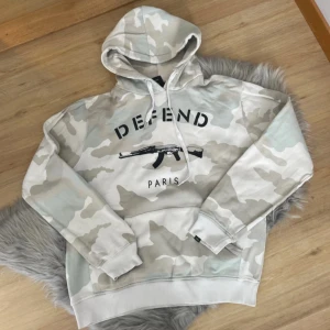 Camouflage hoodie från Defend Paris - Säljer en cool hoodie från Defend Paris i ett stilrent camouflage-mönster. Tröjan har en stor huva med dragsko och en tryckt design på framsidan. Perfekt för en avslappnad stil.
