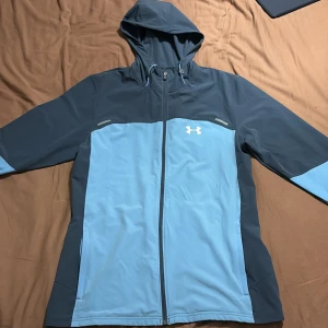 Blå och grå vindjacka från Under Armour - Säljer en snygg blå och grå vindjacka från Under Armour. Jackan har en dragkedja framtill och en praktisk huva. Perfekt för utomhusaktiviteter med sin sportiga design och bekväma passform. Pris kan diskuteras 