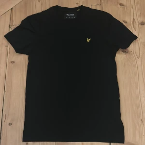 Svart t-shirt från Lyle & Scott - Säljer en stilren svart t-shirt från Lyle & Scott med deras ikoniska logga broderad i gult på bröstet. Perfekt för en casual look. T-shirten är kortärmad och gjord i mjukt bomullsmaterial.