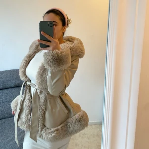 Beige jacka med pälsdetaljer - Snygg beige jacka med fluffiga pälsdetaljer på huva, ärmslut och nederkant. Jackan har ett bälte i midjan för en figursydd look. Perfekt för kyliga dagar när du vill hålla stilen.
