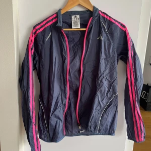 Grå vindjacka från Adidas - Säljer en grå vindjacka från Adidas med rosa detaljer och dragkedja. Jackan har långa ärmar och de klassiska Adidas-ränderna i rosa längs ärmarna. Hade gärna behållt men är något liten på mig, passar en strl S!