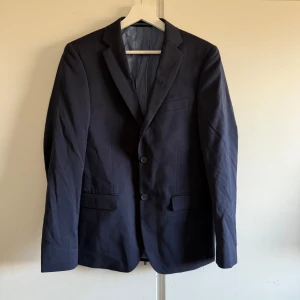 Mörkblå kostym från Dressmann - Säljer en stilren mörkblå kostym från Dressmann. En av knapparna på armen lite lös annars är den i bra skick. Kavajen är storlek 46R i slim fit och byxorna är 31/32 slim fit. En slips följer med på köpet också. 