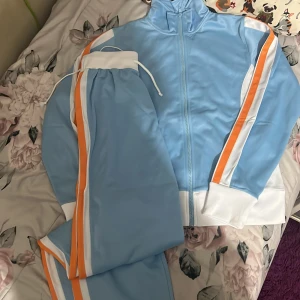 Blå träningsjacka med dragkedja - Snygg ljusblå träningsjacka med dragkedja och vita samt orange ränder längs ärmarna. Jackan har en hög krage och ribbade muddar. Perfekt för sportiga aktiviteter eller en avslappnad stil.