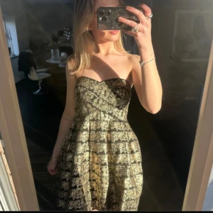 Axelbandslös klänning från Sans Souci - Snygg axelbandslös klänning från Sans Souci i svart och guld med ett vackert spetsmönster. Perfekt för festliga tillfällen med sin eleganta design och glittriga detaljer. Säljer då den var för stor för mig😔