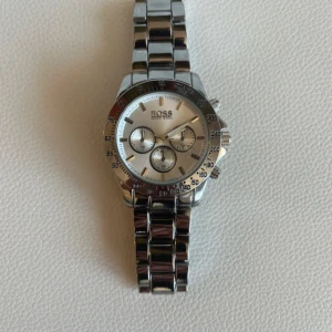 Silverfärgad klocka från Boss - Snygg och stilren silverfärgad klocka från Boss med metallarmband och flera små urtavlor. Perfekt för den som gillar en klassisk och elegant look. Klockan har en tydlig logotyp på urtavlan och baksidan av armbandet. Tillkommer fler länkar.