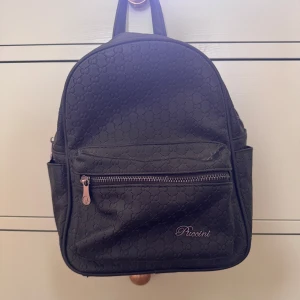 Svart ryggsäck från Piccini - Snygg svart ryggsäck från Piccini med diskret mönster och dragkedja framtill. Väskan har justerbara axelremmar och flera praktiska fack för enkel organisering. Perfekt för både skola och fritid!