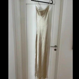 Champagnefärgad sidenklänning - Långklänning i champagnefärgat siden med smala axelband och öppen rygg. Klänningen hardrapering framtill och knytband i ryggen för en justerbar passform och sen liten slits längst ner. Använd en gång