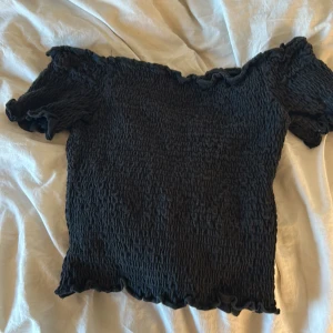 Svart ribbad offshoulder topp - Snygg svart offshoulder topp med ribbad struktur. Perfekt för en stilren look. Toppen har korta ärmar och en elastisk passform som ger en smickrande siluett.