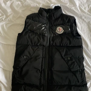 Svart dunväst från Moncler - Snygg svart dunväst från Moncler med dragkedja och broderad logga på bröstet. Västen har praktiska fickor med knappar och är perfekt för kyligare dagar.