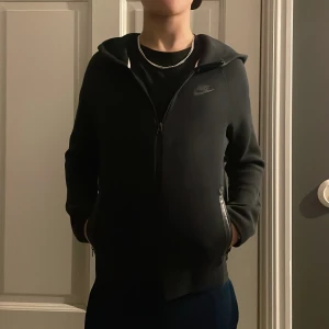 Svart Nike tech från Nike - Säljer en stilren svart Nike tech från Nike med dragkedja och huva. Den passar till mycket och är i bra skick