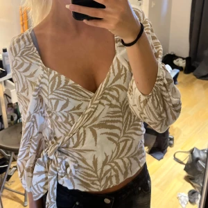 Beige och vit omlottblus  - Mönstrad omlottblus i beige och vitt. Blusen har trekvartsärmar, v-ringning och knyt i sidan. Storlek 40 men i och med att den är justerbar passar den även mig som annars bär xs/s. Sparsamt använd. Säljer för 100kr.