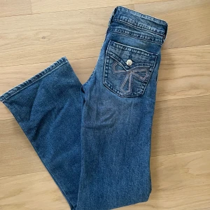 Blå bootcut jeans med broderad ficka - Snygga blå jeans med bootcut passform och cool broderad detalj på bakfickan. Klassisk femficksmodell med knappstängning och slitna detaljer för en avslappnad look. Perfekta till sneakers eller boots. 