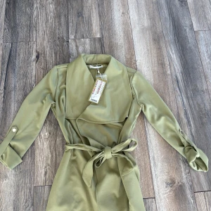 Olivgrön trenchcoat med bälte - Snygg olivgrön trenchcoat från New Yorker med bred krage och knytbälte i midjan. Jackan har långa ärmar med knappdetalj vid mudden och en omlott-design framtill. Perfekt för dig som vill ha en stilren och trendig look.