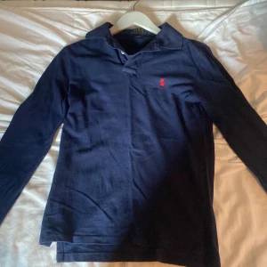 Marinblå långärmad pikétröja Polo Ralph Lauren XS - Snygg marinblå långärmad pikétröja från Polo Ralph Lauren i slim fit. Tröjan har klassisk krage, knäppning vid halsen och den ikoniska röda broderade loggan på bröstet. Tillverkad i mjuk bomull som är skön mot huden.