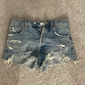 Blå slitna jeansshorts med fransar - Riktigt snygga blå jeansshorts med lägre midja och råa, fransiga kanter. De har flera slitningar och hål både fram och bak för en cool, edgy vibe. Klassisk femficksmodell med knapp. Perfekta för sommarens alla äventyr.