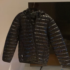 Svart pufferjacka från Everest - Snygg svart pufferjacka från Everest i storlek 170/176. Jackan har glansig yta, horisontella quiltade sömmar, huva och dragkedja framtill. Perfekt för kalla dagar och riktigt lätt att bära. Klassisk look som funkar till det mesta.