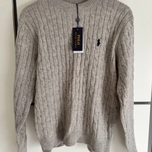  tröja Polo Ralph Lauren - Kabelstickad grå tröja från Polo Ralph Lauren med klassisk rund hals och broderad svart logga på bröstet. Tröjan har lång ärm och ribbade muddar. Perfekt för dig som gillar stilren och tidlös design.