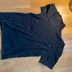 Svart t-shirt från Calvin Klein - Svart t-shirt från Calvin Klein med klassisk passform och diskret CK-logga broderad på bröstet. Tillverkad i mjuk bomull och har rund halsringning samt korta ärmar. Perfekt för en clean och stilren look.