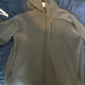 Grå hoodiejacka från Sail Racing - Mörkgrå hoodiejacka från Sail Racing med hel dragkedja framtill och bröstficka med dragkedja. Jackan har en stilren look, huva och diskret logga på bröstet. Perfekt för dig som gillar sportig och clean stil.