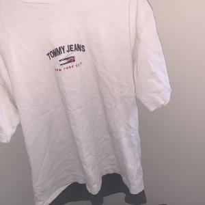 Vit t-shirt Tommy Jeans S - Vit t-shirt från Tommy Jeans med broderad logga och text 'New York City' på bröstet. Klassisk passform, kortärmad och med svart kant längst ner. Tillverkad i mjuk bomull, perfekt för en clean och stilren look. Pris kan diskuteras vid snabb affär