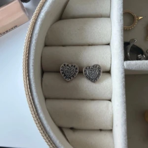 Pandora hjärtformade örhängen i silver - Säljer ett par snygga hjärtformade örhängen från Pandora i äkta silver. De har glittriga stenar i mitten och Pandora-loggan runt kanten. Perfekta för dig som gillar detaljer och vill ha något som sticker ut lite extra.