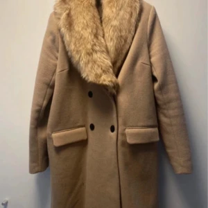 Beige kappa med fuskpäls från Ellos - Snygg beige kappa från Ellos med bred krage i fuskpäls. Dubbelknäppt framtill med svarta knappar och två stora fickor. Långärmad och rak modell, perfekt för kalla dagar. Materialet känns mjukt och värmande.