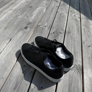 Svarta sneakers från Björn Borg - Stilrena svarta sneakers från Björn Borg i skinn med klassisk vit platt sula. Skorna har rund tå, svarta snören och diskret Björn Borg logga på sidan. Skorna har även aldrig använts.
