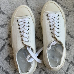 Vita sneakers från Jil Sander - Stilrena vita sneakers från Jil Sander med canvasöverdel och robust, krämfärgad gummisula. Klassisk rund tå, vita snören och diskret logga på både sula och häl. Perfekta för en clean och minimalistisk look.