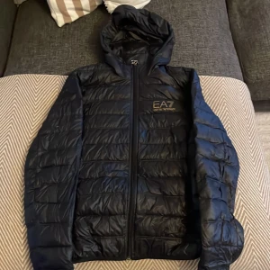 Svart dunjacka från EA7 Emporio Armani - Säljer en svart dunjacka från EA7 Emporio Armani med huva och dragkedja. Jackan har quiltad design, guldig logga på bröstet och sitter snyggt med normal passform. Perfekt för kyliga dagar och enkel att matcha med allt.