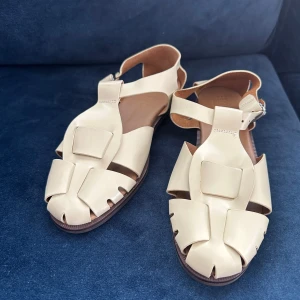 Beige sandaler från Hereu i skinn - Snygga beige sandaler från Hereu i äkta skinn med flätad design och justerbart spänne vid ankeln. Platt sula och öppen tå ger en chill vibe, perfekt för varma dagar. Tillverkade i Spanien och riktigt stilrena till både jeans och klänning.