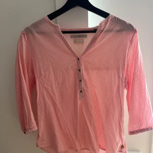 Maison Scotch rosa randig blus - Säljer en söt rosa och vit randig blus från Maison Scotch med v-ringning och dekorativa knappar framtill. Blusen har trekvartsärm och fina spetsdetaljer vid halsringningen och ärmsluten. Perfekt för vår och sommar, superlätt och skön i bomull.