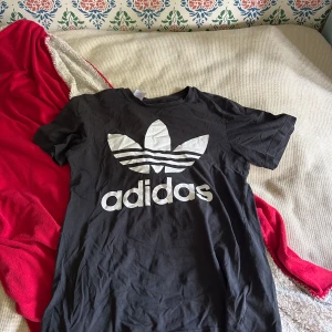 Svart Adidas t-shirt med logga - Svart t-shirt från Adidas med stor vit treklöver-logga och Adidas-tryck på bröstet. Klassisk rund hals och korta ärmar. Tillverkad i mjuk bomull som är skön mot huden. Perfekt för dig som gillar sportig och enkel stil.