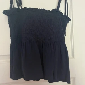 Mörkblå smockad linnetopp med knyt - Supersöt mörkblå linnetopp med smockad överdel och volangkant upptill. Toppen har tunna axelband som knyts på axlarna för en personlig touch. Den är luftig och har en lätt rynkad nederdel som ger en chill vibe.