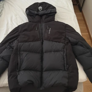 Svart Sail Antarctica pufferjacka - Säljer en svart pufferjacka från Sail racing Expedition. Jackan har huva, dragkedja framtill och flera fickor med dragkedja. Den är vadderad och har en snygg badge på ärmen. Perfekt för kalla dagar när du vill hålla stilen och värmen.