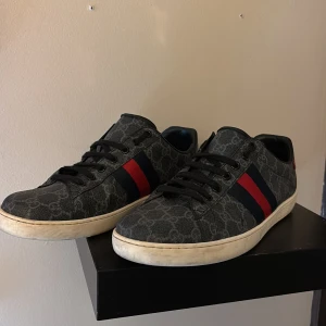 Gucci ace  - Snygga Gucci sneakers i mörkgrått med klassiskt GG-mönster och svarta snören. Sidorna har röda och marinblå ränder, och hälarna är metallicröd och metallicblå med Gucci-logga. Yttermaterialet är canvas och detaljer i skinn. Platt sula med unik design under. Smutsen går bort med lite vatten finns också en skada på vänster skon som syns på bild. BYTEN KAN VARA INTRESSENT TYP SOM MONCLER. Kom med bud. Har box o allt sånt