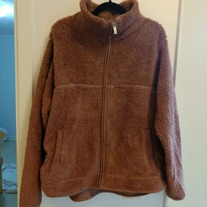 Brun teddyjacka med dragkedja - Mysig brun teddyjacka med hög krage och hel dragkedja framtill. Jackan har en loose passform och är tillverkad i fluffigt teddy-material som håller dig varm och bekväm. Perfekt för kyliga dagar när du vill ha en avslappnad och trendig look.