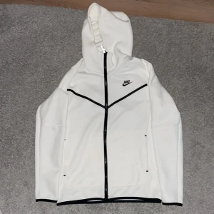 Vit Nike tech med huva - (Storlek XS men passar S också) Aldrig använd Nike tech jag har bara testat den men den passade inte mig. Vit Nike tech med svarta detaljer och hel dragkedja. Tröjan har huva, lång ärm och praktiska fickor, samt en extra ficka med dragkedja på ena ärmen. Perfekt för dig som gillar sportig och clean stil.