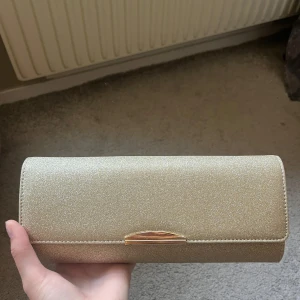 Glittrig guldig kuvertväska Anna Field - Snygg kuvertväska från Anna Field i glittrigt guld med stilren metallstängning och kedja i matchande färg. Den är 27x11,5cm och använd två gånger men finns inga tecken på användning