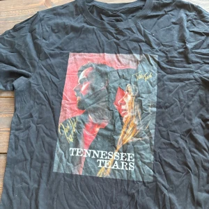 Signerad Svart Tennessee Tears t-shirt  - Signerad Svart t-shirt med färgglatt Tennessee Tears-tryck framtill. Motiv med två personer och autograf-liknande detaljer i guld. Klassisk rund hals och korta ärmar. Skön och mjuk bomullskänsla, perfekt för dig som gillar bandmerch och vill sticka ut.