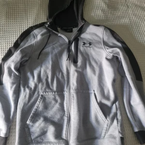 Grå hoodiejacka från Under Armour - Grå hoodiejacka från Under Armour med detaljer på axlar och huva. Jackan har dragkedja framtill, praktiska fickor och en bröstficka med dragkedja. 