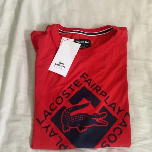 Röd Lacoste Sport t-shirt med tryck - Röd t-shirt från Lacoste Sport med stort mörkblått tryck framtill och klassisk rund hals. Snygg detalj med randig insida i halsen. Perfekt för dig som gillar sportig och stilren design.