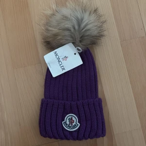 Lila mössa med pälsboll inspirerad av Moncler - Superfin lila ribbstickad mössa med stor fluffig pälsboll upptill. Perfekt accessoar för kalla dagar och ger en snygg touch till din vinterstil.