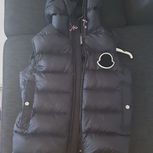 Moncler Vanoise Gilet  - Svart dunväst från Moncler, modell Vanoise Gilet. Västen har en hög krage, huva med snörning och två fickor med tryckknappar. Moncler-logga på bröstet och dragkedja framtill. Priset kan diskuteras.