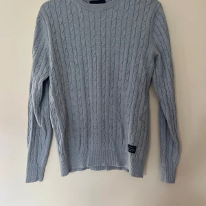 Ljusgrå kabelstickad tröja Bondelid - Kabelstickad tröja från Bondelid i ljusgrått med rund halsringning. Klassisk modell med ribbade muddar vid ärmslut och nederkant. Perfekt för lager-på-lager och chill stil. Snygg detalj med diskret logolapp nertill.