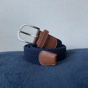 Blå flätad bälte med bruna detaljer - Snyggt flätat bälte i marinblått med bruna skinn-detaljer och silverfärgat spänne. Perfekt för att ge din outfit en stilren touch. Bältet har vita sömmar på skinnet och en klassisk form som passar till jeans eller chinos. Bältet är 110cm långt