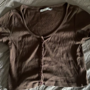 Brun ribbad topp med knappar 157 - Kortärmad brun ribbad topp från 157 i storlek XS. Toppen har croppad passform, djup rundringning och knappar framtill. Perfekt att styla med höga jeans eller kjol för en chill look.
