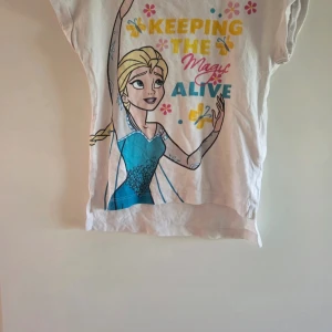 Frozen t-shirt med motiv av Elsa - Vit kortärmad t-shirt från Disney med färgglatt tryck av Elsa från Frozen och texten 'Keeping the magic alive'. T-shirten har rosa och gula blommor, blå detaljer och en rosa kant vid halsen. Tillverkad i mjuk bomull, perfekt för dig som älskar Frost.