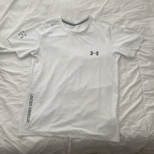 Vit Under Armour t-shirt med reflex - Snygg vit t-shirt från Under Armour med coola reflexdetaljer på bröstet, axeln och ärmen. T-shirten har rund hals, korta ärmar och tryck längs sidan. Perfekt för träning och löpning, tillverkad i lätt polyester som andas.
