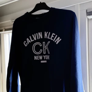 Mörkblå Calvin Klein sweatshirt M - Snygg mörkblå sweatshirt från Calvin Klein Jeans i storlek M. Tröjan har vit CK New York-logga framtill, rund hals och långa ärmar. Mjuk bomullskvalitet och klassisk passform som funkar till jeans eller joggers. Perfekt för dig som gillar stilrena märkesplagg.