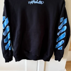  Off-White hoodie  - Säljer en svart hoodie från Off-White Kids med ikoniska blå pilar på ryggen och blått tryck på ärmarna. Tröjan har huva, magficka och ribbade muddar. Materialet är mjuk bomull och polyester, perfekt för dig som gillar streetwear och vill sticka ut med cool design. Till barn som är 10 år gammal ungefär